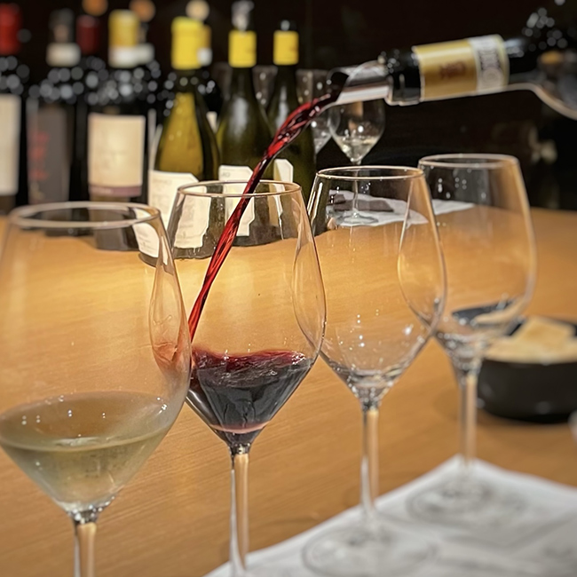 Calice di vino rosso versato con precisione accanto a tre calici vuoti e uno di vino bianco, su bancone elegante con bottiglie sullo sfondo