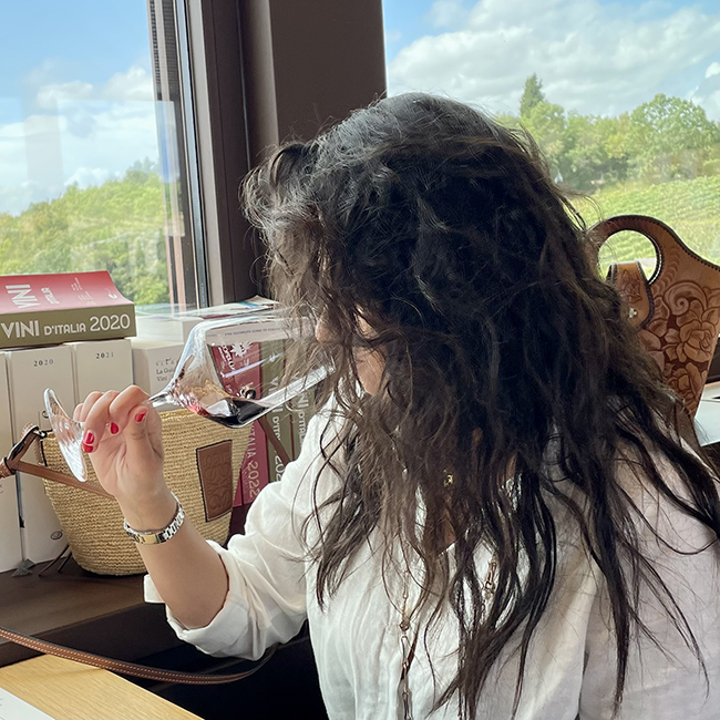 Donna osserva un calice di vino rosso con sguardo concentrato, davanti a una finestra che affaccia sui vigneti