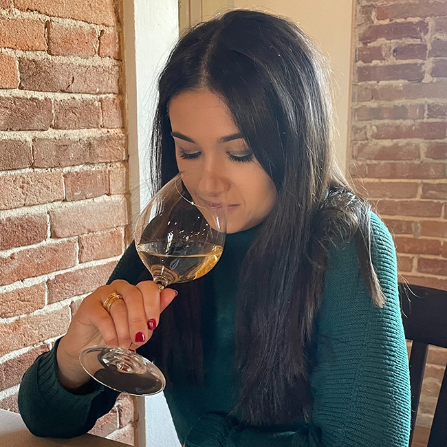 Donna annusa un calice di vino bianco durante una degustazione, seduta accanto a un muro in mattoni