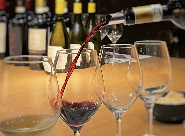 Calice di vino rosso versato con precisione accanto a tre calici vuoti e uno di vino bianco, su bancone elegante con bottiglie sullo sfondo
